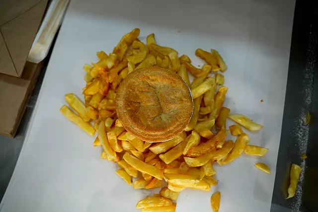 A golden shortcrust pie sitting on a bed of chips, wrapped open on paper
