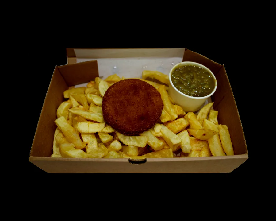 A round breadcrumbed fish cake on thick-cut chips in an open takeaway box, with a pot of mushy peas